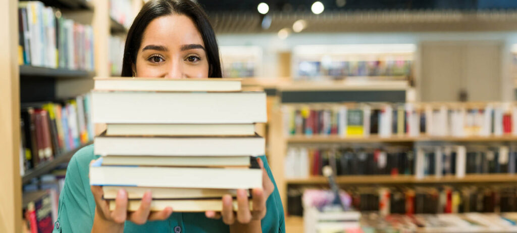 Immagine evocativa per scuola librai. Libraia con il viso parzialmente nascosto da una pila di libri. Sullo sfondo la libreria